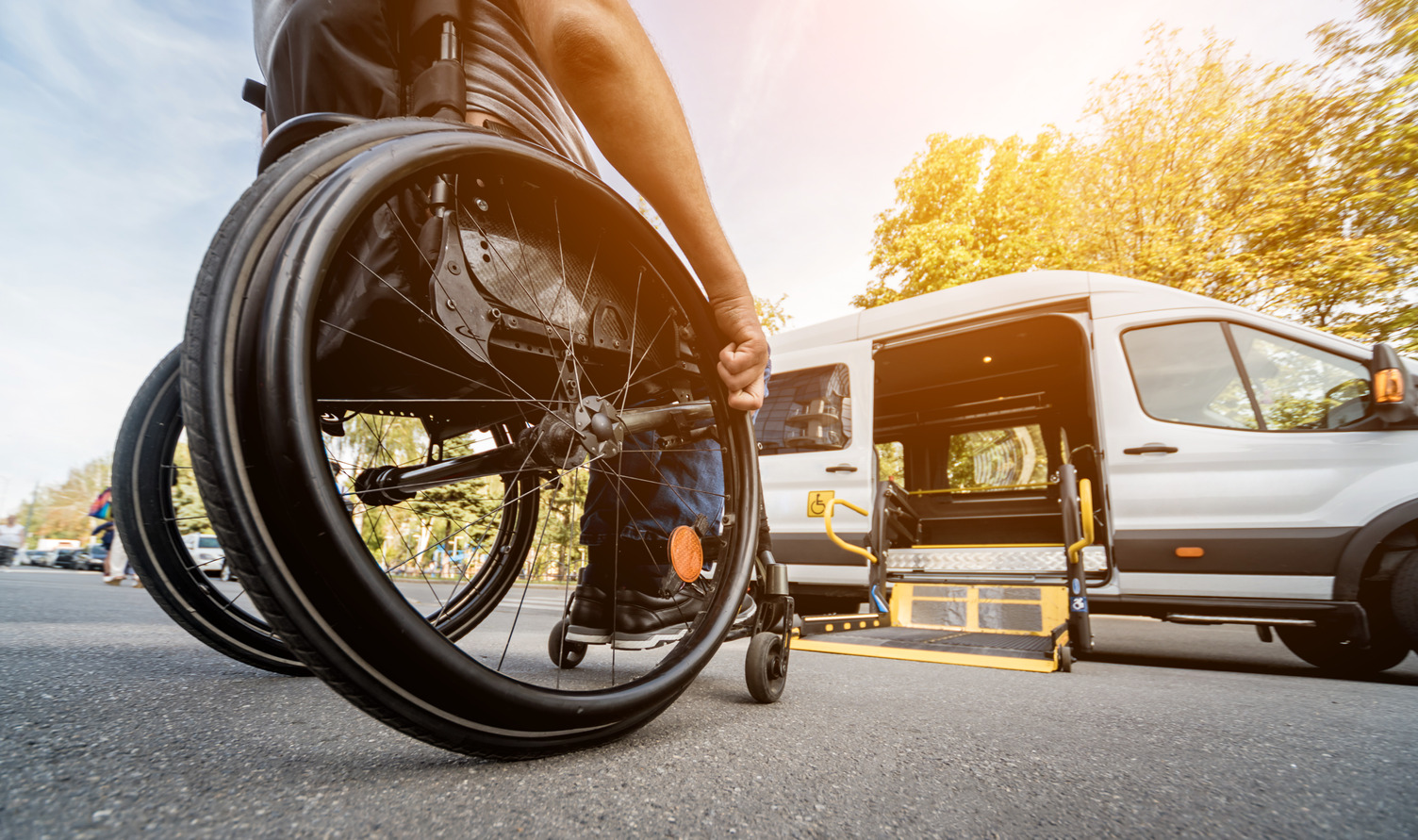 AdobeStock_386147368 A person in a wheelchair moves towards a wheelchair lift on a specialized transit vehicle parked outside during the day.