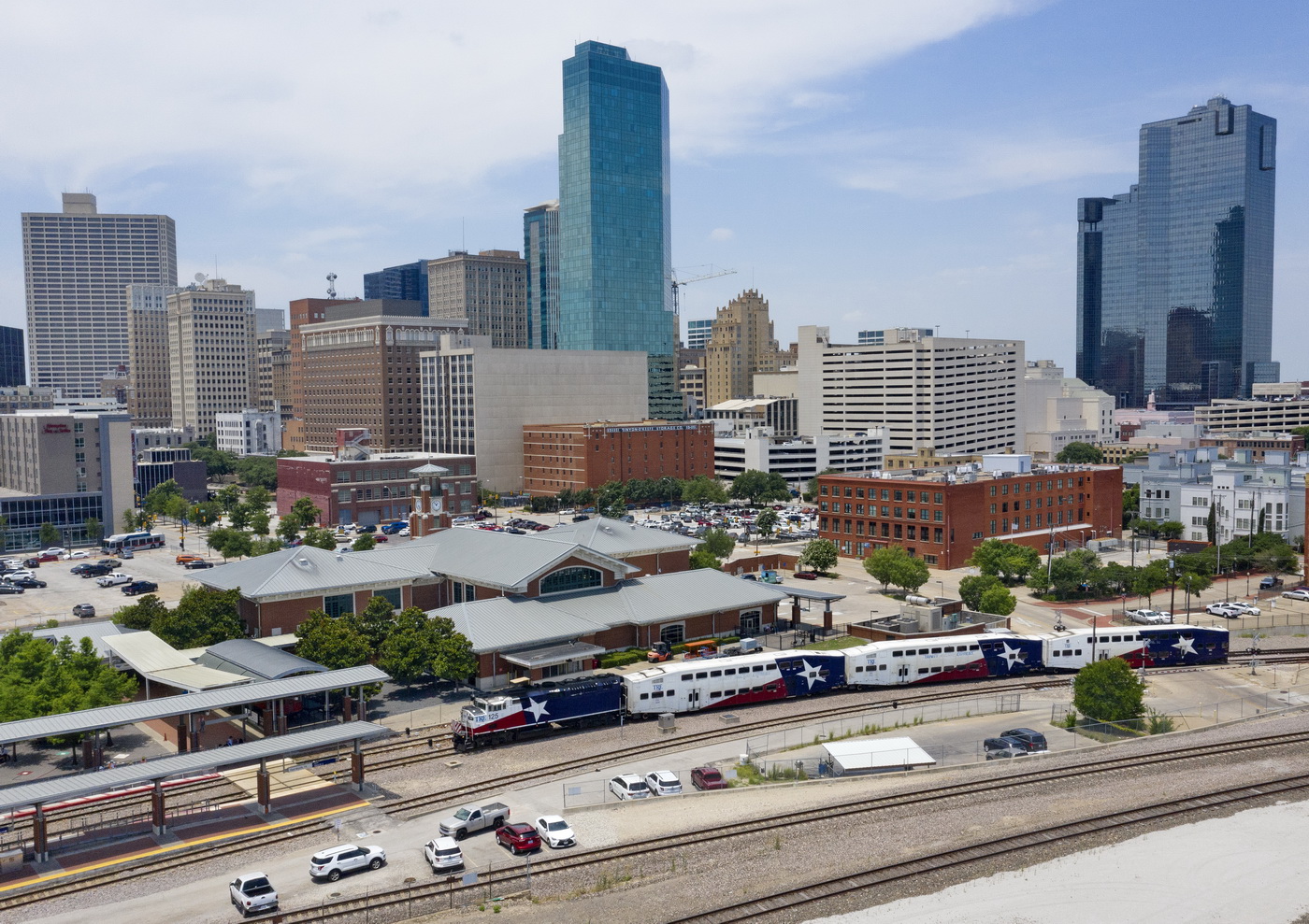 Trinity Metro TRE Passing Fort Worth Central Station A high-angle aerial view of a Trinity Railway Express (TRE) commuter train passing the Fort Worth Central Station on a bright, sunny day. The train consists of a locomotive and several passenger cars, all featuring a distinctive red, white, and blue paint scheme with white stars, resembling the Texas flag. The station itself is a large, low-rise brick building with a multi-gabled gray roof, situated next to the tracks and several parking lots. The background features the dense downtown Fort Worth skyline, prominently showcasing the blue glass Omni Fort Worth Hotel and other modern skyscrapers against a clear sky.