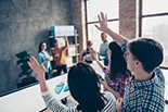 Students raising hands in a classroom to ask presenters a question.